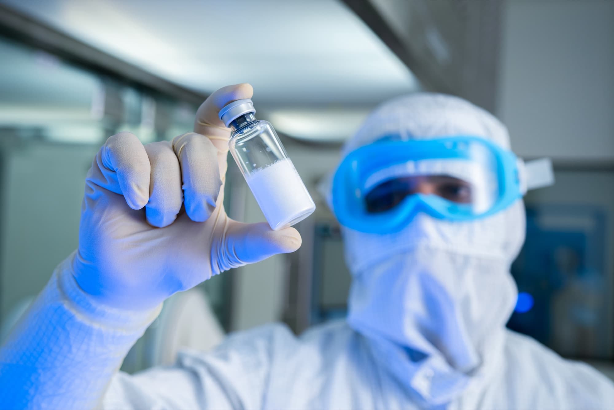 A gloved hand holds up a glass vial of lyophilized white powder for inspection, with a cleanroom-suited researcher in the background.