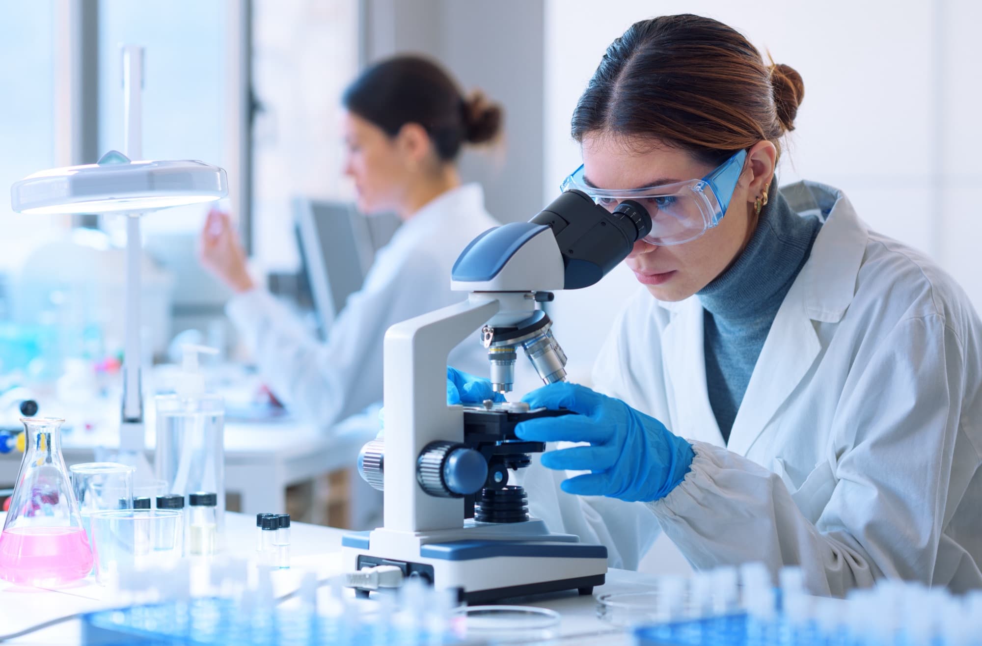 A researcher in safety glasses and a lab coat examines a sample under a microscope, with a colleague working at a lab bench in the background.